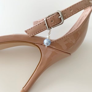 May include: A glossy nude patent leather high-heeled shoe featuring an ankle strap with a silver-toned metal buckle. A small charm consisting of a tiny white bead and a larger, round, light blue pearl-like bead hangs from the buckle hardware.