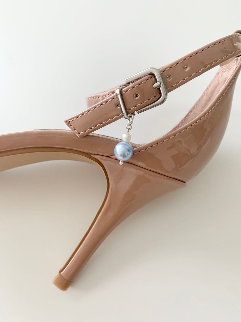 May include: A glossy nude patent leather high-heeled shoe featuring an ankle strap with a silver-toned metal buckle. A small charm consisting of a tiny white bead and a larger, round, light blue pearl-like bead hangs from the buckle hardware.