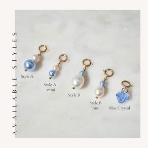 May include: Five gold-toned jewelry charms labeled AVAILABLE STYLES. Style A features a blue bead and white pearl. Style A mini has a smaller blue bead and pearl. Style B shows a large white pearl and blue bead. Style B mini has a smaller pearl and blue bead. Blue Crystal is a faceted blue charm.