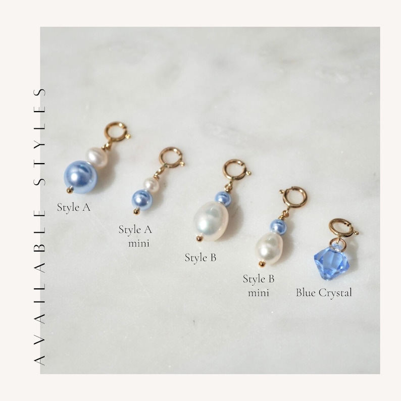 May include: Five gold-toned jewelry charms labeled AVAILABLE STYLES. Style A features a blue bead and white pearl. Style A mini has a smaller blue bead and pearl. Style B shows a large white pearl and blue bead. Style B mini has a smaller pearl and blue bead. Blue Crystal is a faceted blue charm.