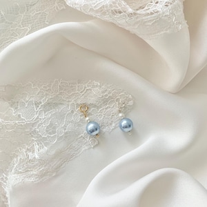 May include: Two small charms featuring a single light blue pearl-like bead and a smaller white bead. One charm has a gold-toned circular clasp, while the other has a silver-toned circular clasp. The charms are positioned on a white fabric surface.