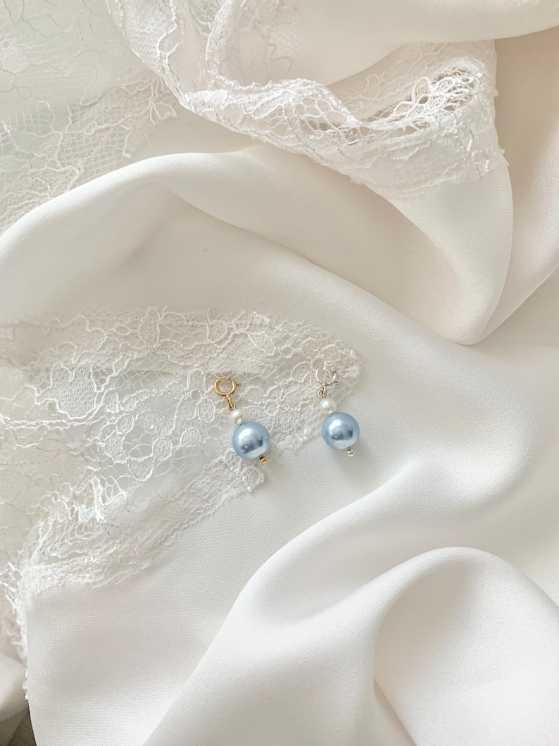 May include: Two small charms featuring a single light blue pearl-like bead and a smaller white bead. One charm has a gold-toned circular clasp, while the other has a silver-toned circular clasp. The charms are positioned on a white fabric surface.