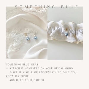 May include: A charm featuring a light blue pearl and a smaller white pearl on a gold-toned clasp. The text SOMETHING BLUE is at the top. Below, the text reads: SOMETHING BLUE IDEAS: - ATTACH IT ANYWHERE ON YOUR BRIDAL GOWN MAKE IT VISIBLE OR UNDERNEATH SO ONLY YOU KNOW ITS THERE! - ADD IT TO YOUR GARTER.