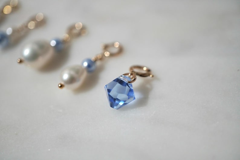 May include: A collection of jewelry charms featuring a faceted light blue crystal pendant attached to a gold-toned jump ring, alongside other charms consisting of white teardrop-shaped pearls and small round light blue beads on gold-toned rings.