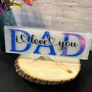 May include: A white rectangular sign with the word "DAD" in blue letters and the text "i love you" in black cursive with hearts. The sign is on a wooden stand.