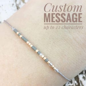 May include: A gray and gold beaded bracelet with a custom message option. The bracelet features a series of gray, gold, and silver beads strung on a thin, light gray cord. Text on the image reads "Custom MESSAGE up to 22 characters."
