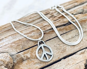 May include: A silver peace sign pendant necklace on a silver snake chain. The necklace is displayed on a piece of driftwood.