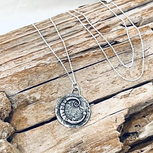 May include: A silver necklace with a round pendant featuring a stylized letter C. The pendant is textured and has a hammered finish. The necklace is on a wooden background.