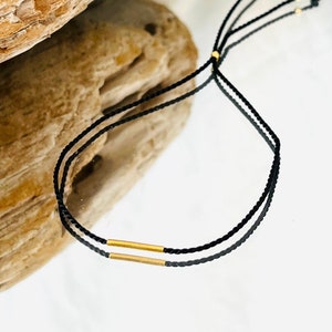 May include: A black string bracelet with two gold bar accents. The bracelet is adjustable and can be worn on the wrist or ankle.