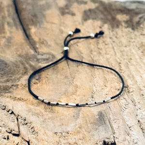 May include: A black cord bracelet with silver-colored rectangular beads. The bracelet has an adjustable sliding knot closure with two small silver beads. The bracelet is displayed on a wooden surface.