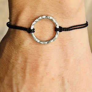 May include: A silver-colored hammered metal circle bracelet strung on black cord. The bracelet is knotted on each side of the circle. The bracelet is worn on a wrist.