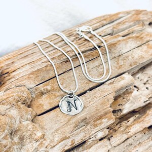 May include: A silver-toned necklace with a pendant featuring the letter "N" in a stylized font. The pendant is circular with a hammered texture. The necklace rests on a piece of weathered wood.