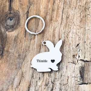 May include: A silver rabbit keychain with a heart cutout and the word "Thimble" engraved on it. The keychain is attached to a silver key ring.