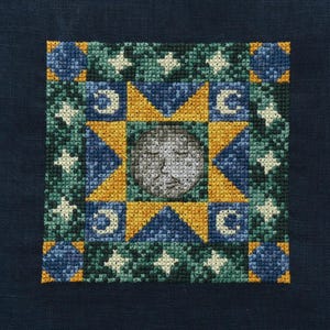 May include: A square, cross-stitched textile featuring a celestial design. The central image is a gray face surrounded by yellow triangles, blue squares, white stars, and crescent moons. The background is a dark blue fabric.