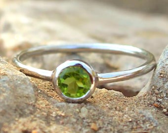 May include: A silver ring featuring a single, round, faceted, lime-green peridot gemstone set in a smooth, circular bezel setting. The band of the ring is thin, polished, and metallic, showcasing a simple design with a vibrant green stone.