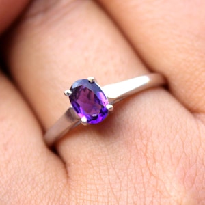 May include: A silver-toned ring featuring a single oval-cut purple gemstone set in a four-prong setting. The band is smooth and polished, tapering slightly as it reaches the central stone, creating a clean and simple design for this piece of jewellery.