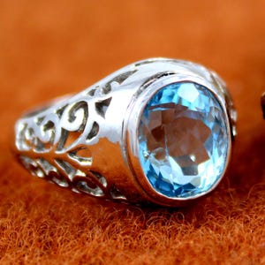 Könnte beinhalten: Ein silberner Ring mit einem ovalen, hellblauen Edelstein. Der Ring hat ein dekoratives, durchbrochenes Design an den Seiten. Der Edelstein ist in einem polierten Silberband gefasst, was einen klassischen und eleganten Look erzeugt. Der Ring liegt auf einer strukturierten, orangebraunen Oberfläche.