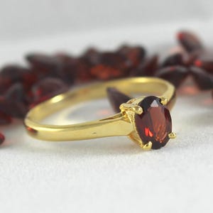 May include: A gold ring with a large, oval-shaped, deep red gemstone. The ring's band is a simple, polished gold, and the gemstone is set with small prongs. The background features scattered red gemstones.