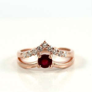May include: Rose gold ring featuring a round, deep red gemstone at the center. The ring has a double band design with a V-shaped top adorned with small, clear accent stones. The ring is set against a white background.