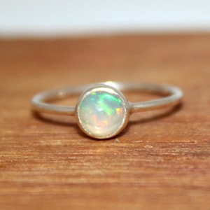 May include: A silver ring featuring a round, bezel-set opal gemstone. The opal displays iridescent flashes of green, orange, and yellow light against a milky white base. The band is thin, smooth, and polished, creating a simple and clean design that highlights the natural play of colour within the central stone.