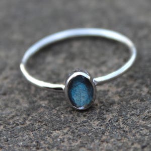 Può includere: Un sottile anello in argento con una piccola pietra preziosa di labradorite di forma ovale. La pietra presenta un riflesso blu iridescente incastonato in una montatura in metallo liscio e lucido. Il gambo è sottile, circolare e uniforme.
