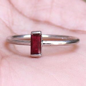 May include: A silver ring featuring a rectangular, deep red gemstone. The gemstone is set horizontally across the band. The ring has a simple, elegant design, suitable for everyday wear. The band appears to be made of a polished metal.