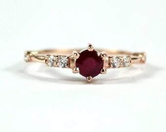 May include: A rose gold ring featuring a central, round, deep red ruby. The band is adorned with small, clear accent stones and intricate detailing. The ring is set against a white backdrop, showcasing its elegant design.