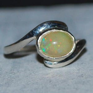 Puede incluir: Un anillo de plata con una piedra de ópalo ovalada. El ópalo tiene un color base amarillo pálido con destellos verdes y rojos. La banda del anillo se curva alrededor de la piedra, creando un diseño único y elegante.