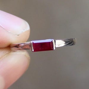 May include: A silver ring featuring a rectangular, deep red gemstone. The band is thin and polished, and the gemstone is set horizontally. The ring is held between fingers against a blurred background.