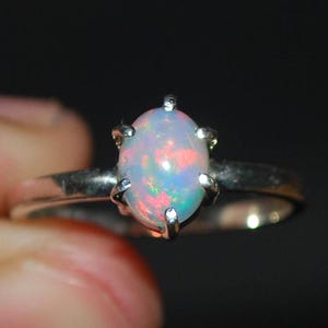 Puede incluir: Un anillo de plata con una piedra de ópalo ovalada. El ópalo muestra colores rosa, azul y blanco, engastado en una montura de cuatro garras. La banda del anillo es de plata pulida.