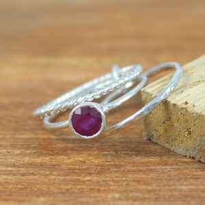 Peut inclure: Trois bagues en argent avec une pierre pr&eacute;cieuse rouge au centre. Les bagues sont empil&eacute;es les unes sur les autres. La bague avec la pierre pr&eacute;cieuse est une simple bande, tandis que les deux autres bagues ont un design torsad&eacute;.