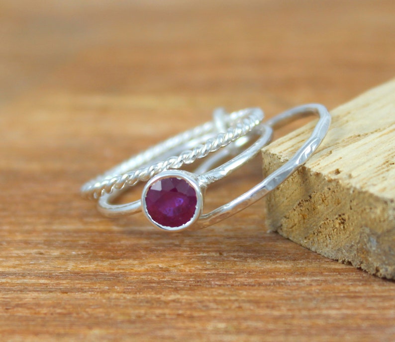 Peut inclure: Trois bagues en argent avec une pierre pr&eacute;cieuse rouge au centre. Les bagues sont empil&eacute;es les unes sur les autres. La bague avec la pierre pr&eacute;cieuse est une simple bande, tandis que les deux autres bagues ont un design torsad&eacute;.