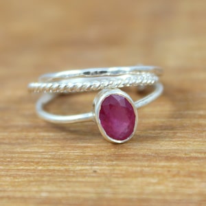 Puede incluir: Un conjunto de anillos de plata con tres bandas. El anillo principal presenta una gema ovalada de color rojo intenso. Las otras dos bandas tienen un dise&ntilde;o de cuerda trenzada y un acabado liso y pulido. Los anillos se exhiben sobre una superficie de madera.