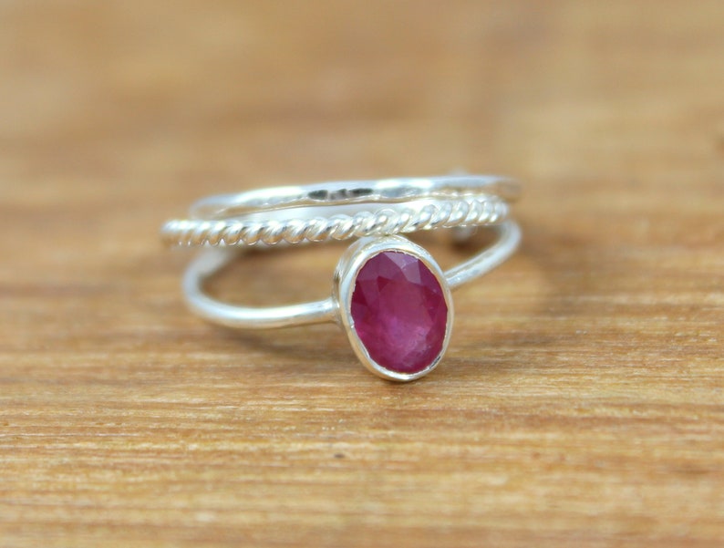 Puede incluir: Un conjunto de anillos de plata con tres bandas. El anillo principal presenta una gema ovalada de color rojo intenso. Las otras dos bandas tienen un dise&ntilde;o de cuerda trenzada y un acabado liso y pulido. Los anillos se exhiben sobre una superficie de madera.