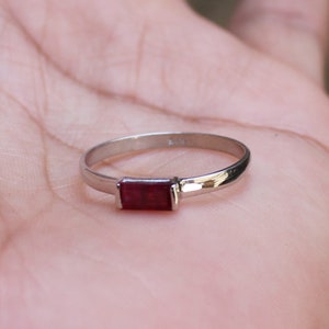 Könnte beinhalten: Ein silberner Ring mit einem rechteckigen, tiefroten Edelstein. Der Ring hat ein dünnes, poliertes Band, und der Edelstein ist in einem schlichten, eleganten Design gefasst. Der Ring wird vor einem hellen Hintergrund gezeigt.