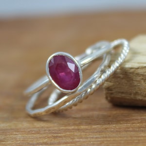 Puede incluir: Un anillo de plata con una gran gema de rub&iacute; de corte ovalado. El anillo tiene un engaste biselado y est&aacute; acompa&ntilde;ado de dos bandas de plata adicionales, una con un dise&ntilde;o de cuerda trenzada. El rub&iacute; es de color rojo intenso.