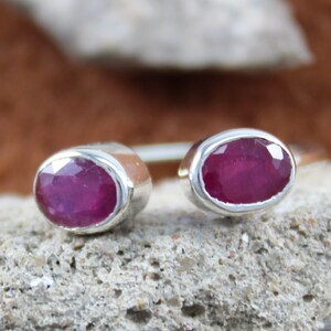 May include: A silver ring with two oval-shaped ruby gemstones. The ring is open and adjustable.