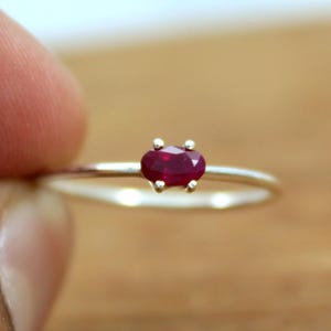 May include: A thin silver ring featuring a small, oval-cut, deep red ruby gemstone set in a four-prong setting. The band is smooth and metallic, showcasing a minimalist design with the vibrant red stone as the central focal point.