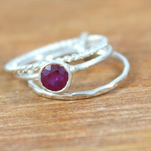 Peut inclure: Trois bagues en argent, dont une avec une pierre pr&eacute;cieuse en rubis rouge, empil&eacute;es sur une surface en bois.