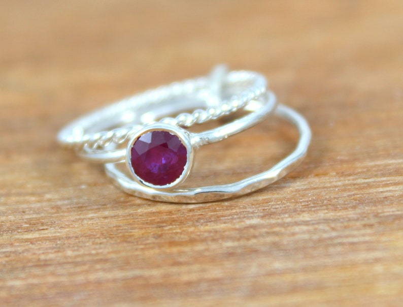 Peut inclure: Trois bagues en argent, dont une avec une pierre pr&eacute;cieuse en rubis rouge, empil&eacute;es sur une surface en bois.