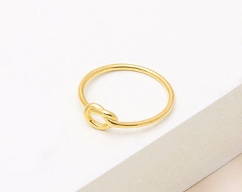 May include: A simple gold ring with a knot design. The ring is on a white surface.