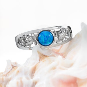 Puede incluir: Un anillo de plata con una piedra preciosa de ópalo azul y un diseño de tres tortugas marinas. El anillo está apoyado en una concha blanca.