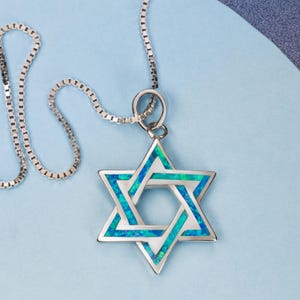 Puede incluir: Un colgante de la Estrella de David en tono plateado con una incrustación iridiscente en tonos azul y verde vibrantes. El dije de estrella de seis puntas cuelga de una delicada cadena de eslabones cuadrados con un marco metálico pulido.