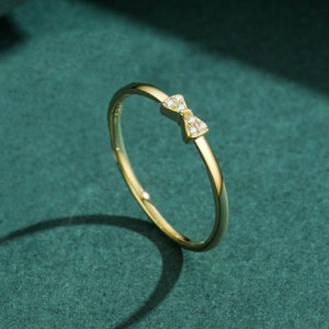 Puede incluir: Un anillo de oro con un pequeño diseño de lazo adornado con piedras de circonita cúbica transparentes.