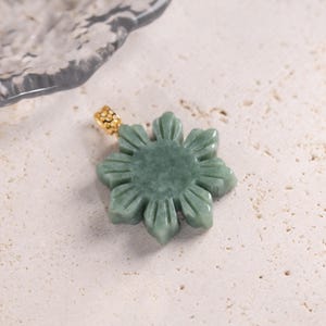 May include: A jade pendant in the shape of a flower with a gold-colored bail. The pendant is a light green color and has detailed carved petals. The bail has a textured design. The pendant is on a light-colored, speckled surface.