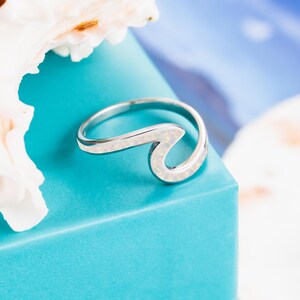 May include: A silver ring with a wave design, featuring a white opal stone. The ring is set against a turquoise background, with a seashell in the upper left corner. The wave design is a popular jewelry motif.