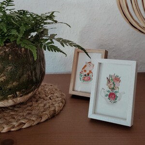 Könnte beinhalten: Zwei gerahmte Aquarellbilder auf einer Holzoberfläche. Das erste Bild zeigt eine Kuh in einer Teetasse. Das zweite Bild zeigt eine Teekanne mit Blumen.