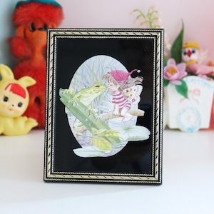 May include: A black picture frame with a gold trim, featuring a whimsical illustration of a girl kissing a frog in a green and brown setting.