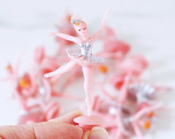 May include: A group of pink ballerina cake toppers with silver and gold accents. The ballerina in the centre is holding her arms out and has a crown on her head.
