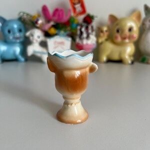 May include: A small, ceramic egg cup with a brown and white glaze. The cup has a scalloped rim and a handle on the side. The cup is sitting on a white surface.